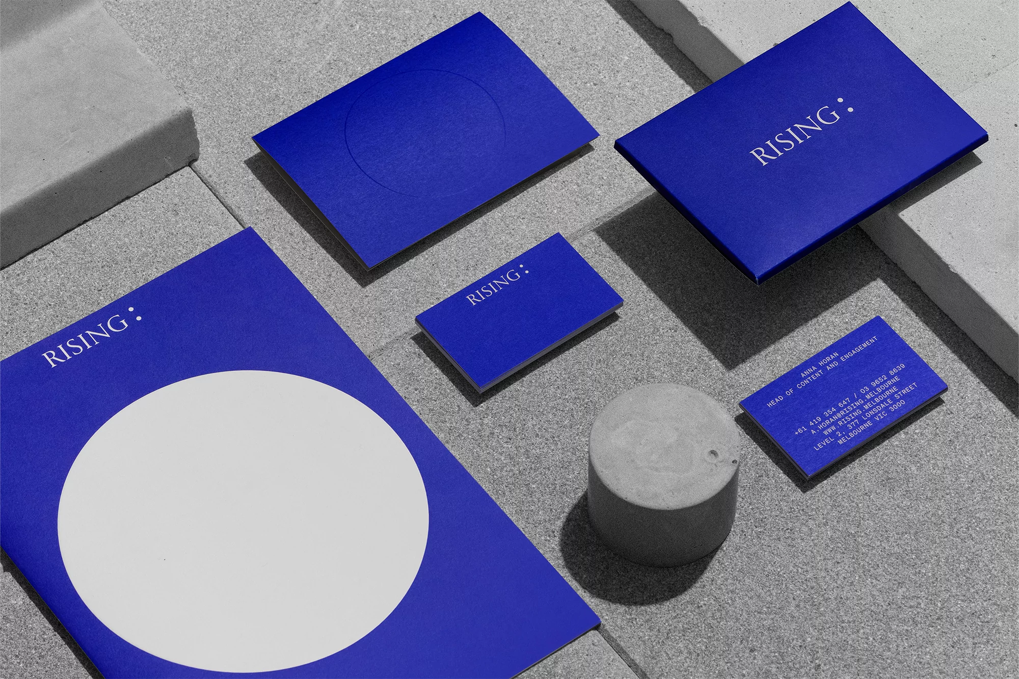 Elevated view of blue marketing materials with 'Rising' text, arranged with a gray concrete cylinder on a textured gray surface.