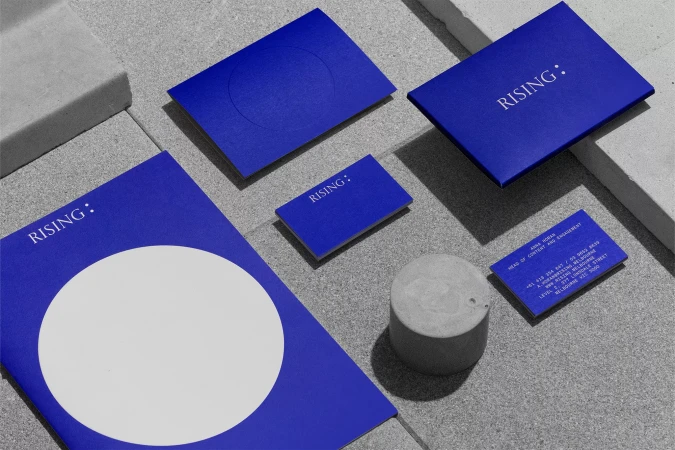 Elevated view of blue marketing materials with 'Rising' text, arranged with a gray concrete cylinder on a textured gray surface.
