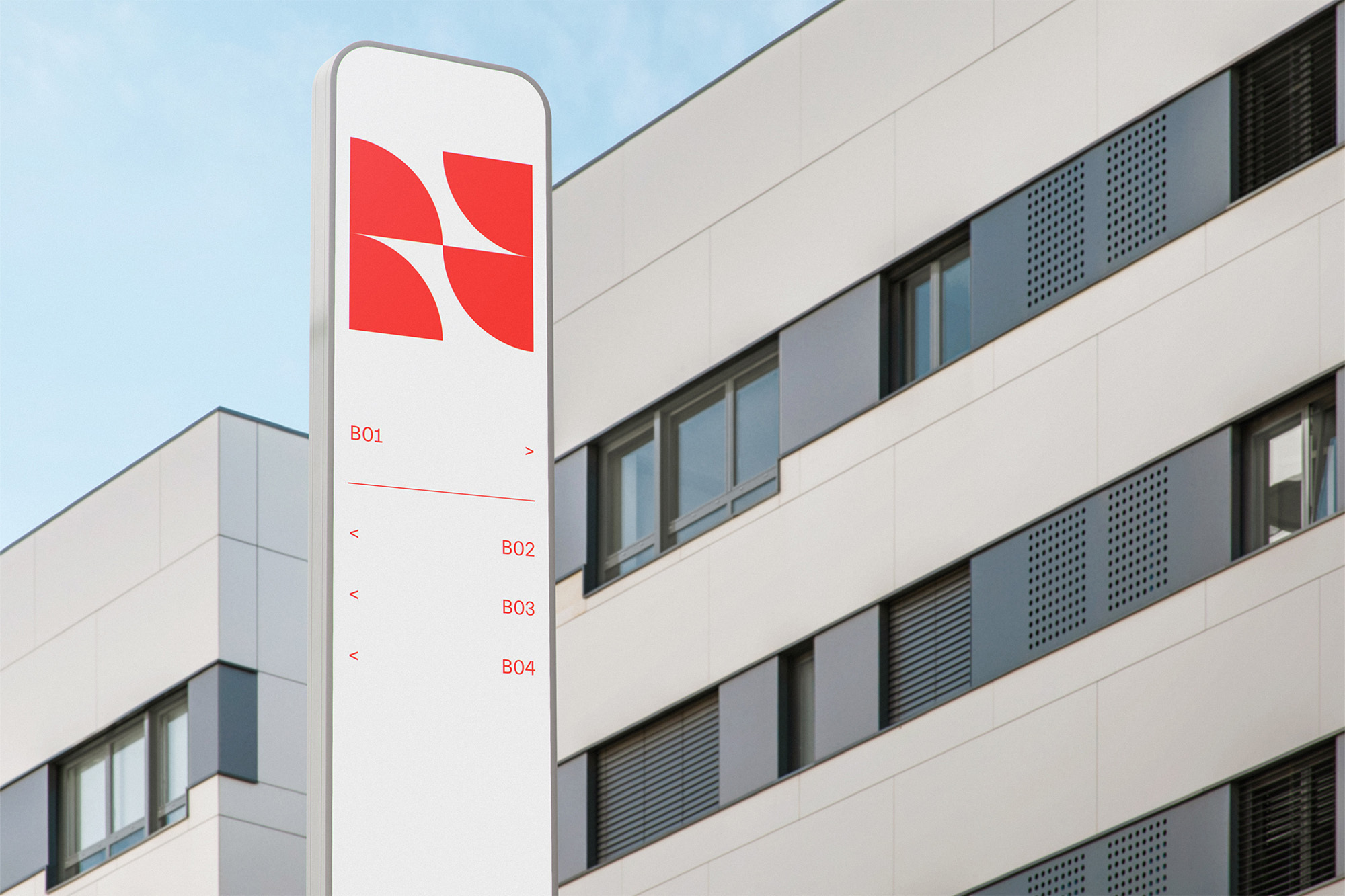 Architectural shot of a sign with a red geometric logo in front of a modern white building with gray window frames and a clear blue sky.