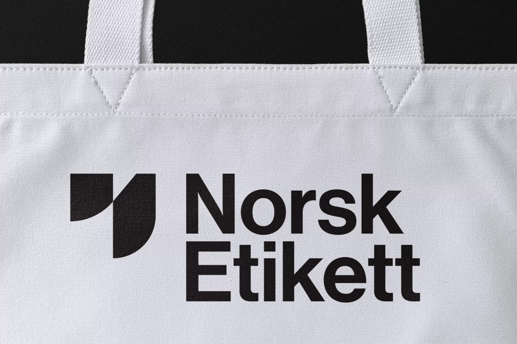 White tote bag featuring the black 'Norsk Etikett' logo. Close-up shot emphasizing branding and minimalist design against a dark background.