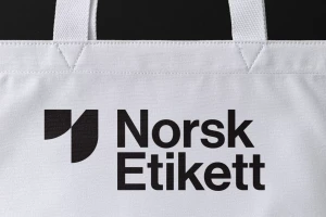 Close-up of a white canvas tote bag featuring the black Norsk Etikett brand logo and graphic mark.