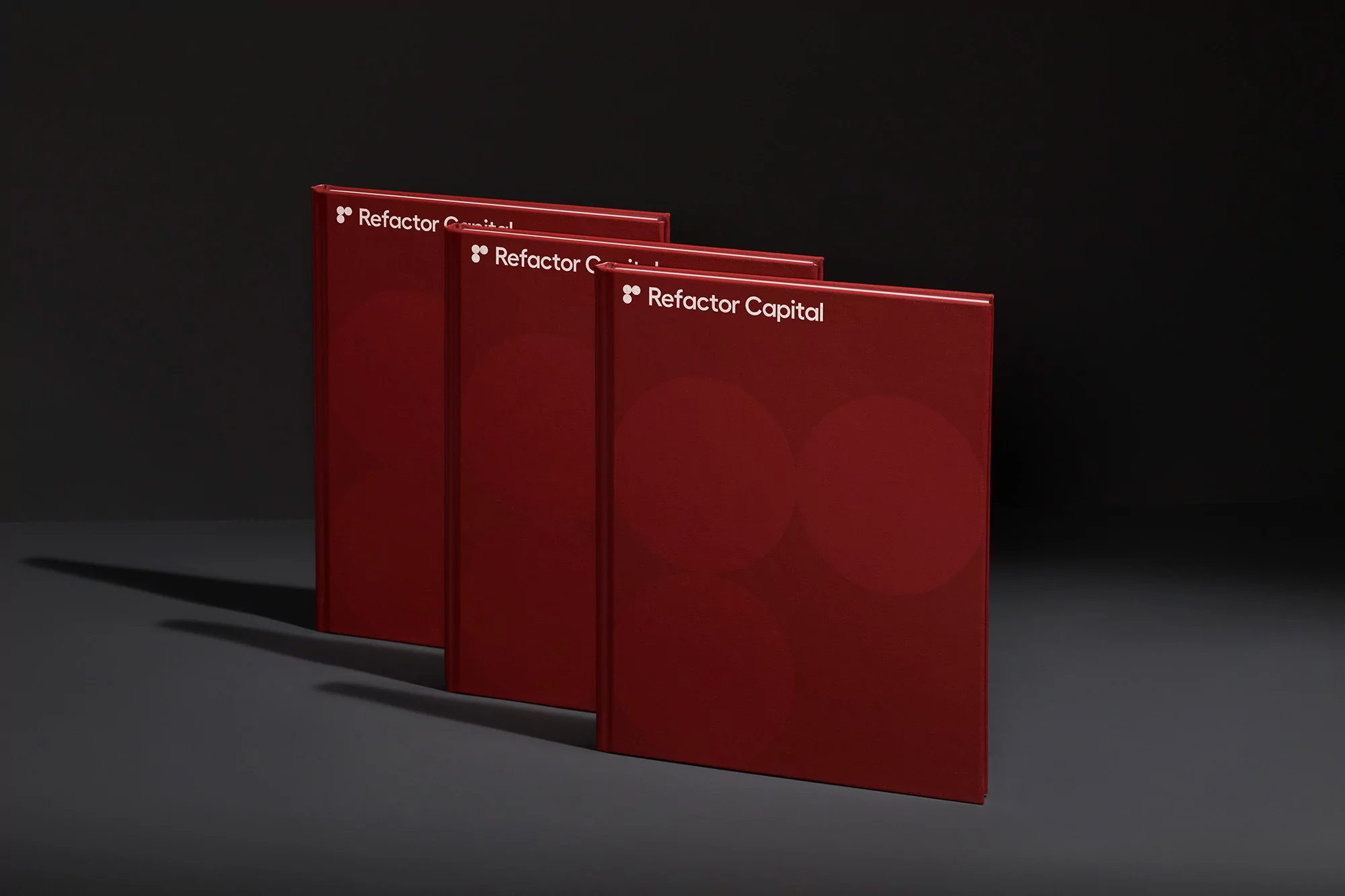 Three red books with the words 'Refactor Capital' stand in a row on a gray surface against a dark gray background in a studio shot.