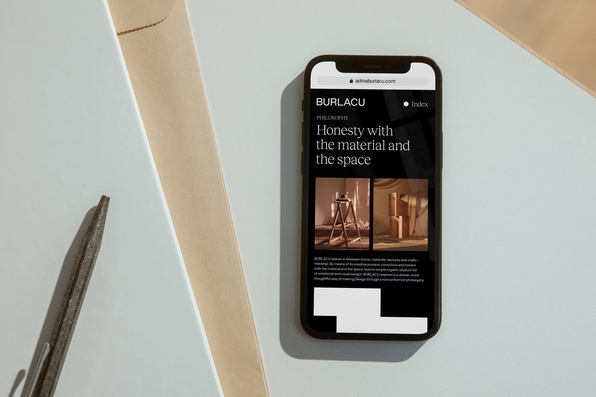 Website for Burlacu Design displayed on a smartphone, next to neutral-toned paper and a gray metal tool on a light blue surface.