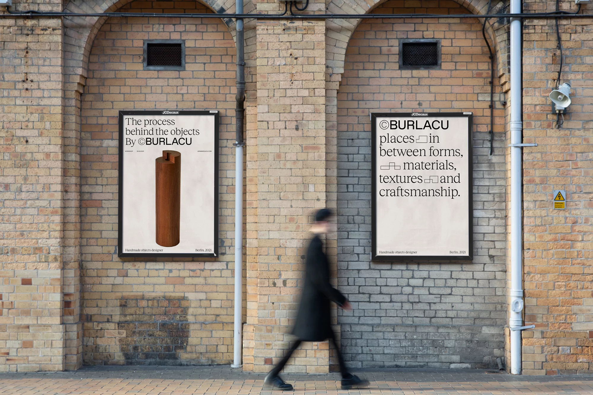 Two Burlacu posters on a brick wall feature graphic design and typography. A blurry person walks by on a sidewalk in an urban setting.