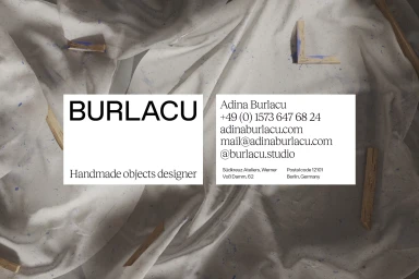 Two business cards with typography resting on draped white fabric with scattered wooden strips and blue paint splatters