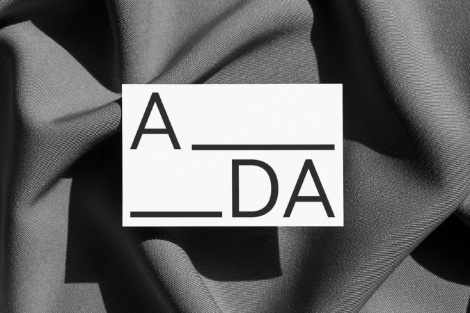 Abstract composition with the text A_DA displayed on a white card, placed on draped gray fabric with soft, natural lighting.