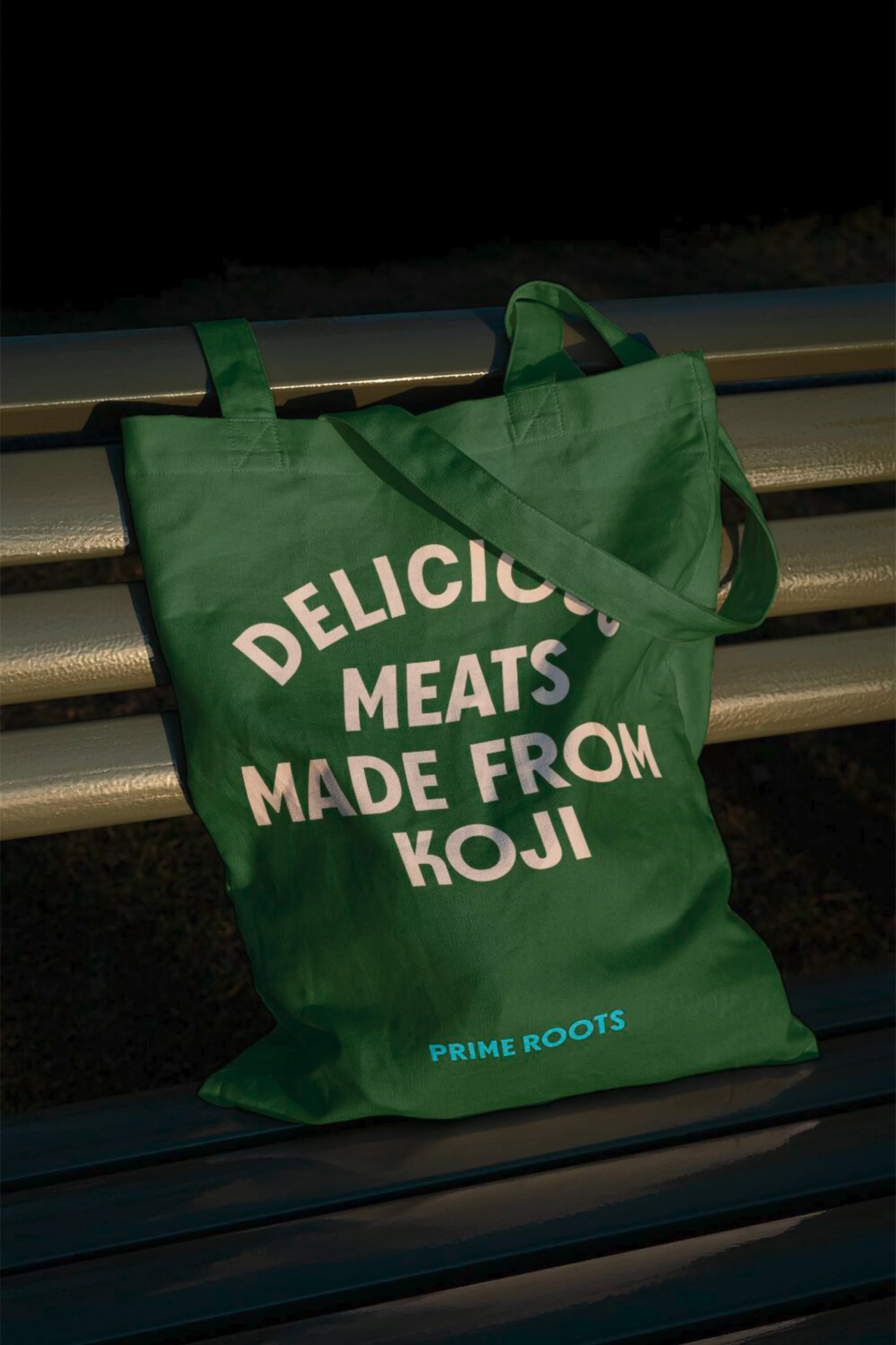 Green tote bag with 'Delicious Meats Made From Koji' text sits on a slatted bench in soft light, showcasing product and sustainable materials.