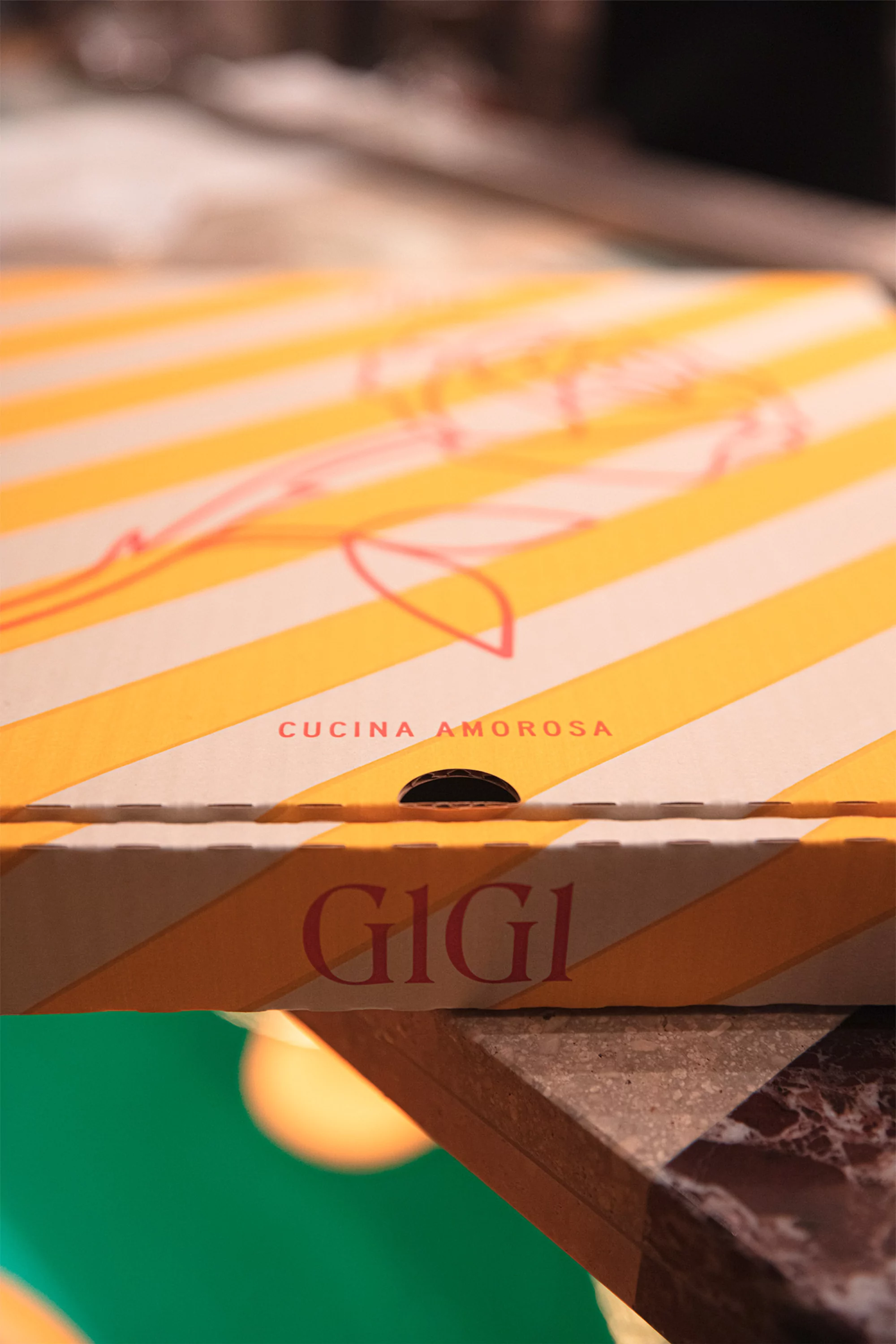 Close-up of a yellow and white striped pizza box with 'Cucina Amorosa' and 'GiGi' printed on it, sitting on a table surface.