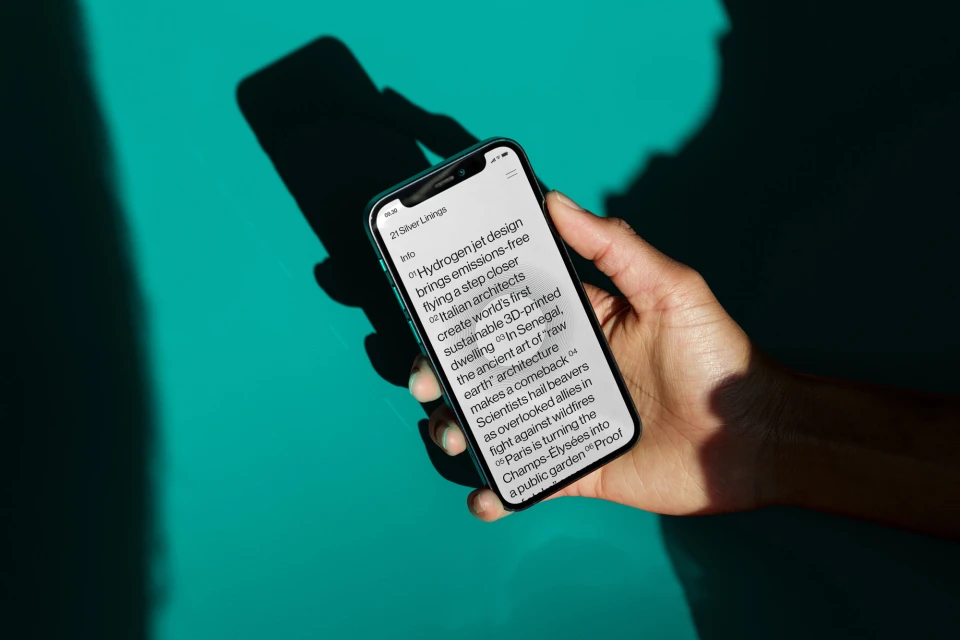 Hand holds smartphone displaying article snippets on design, architecture, and environmental topics against a teal-green background.