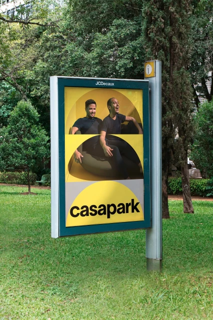 Outdoor advertising billboard featuring two adult men, a yellow graphic design, and the text 'casapark', in a grassy park setting.