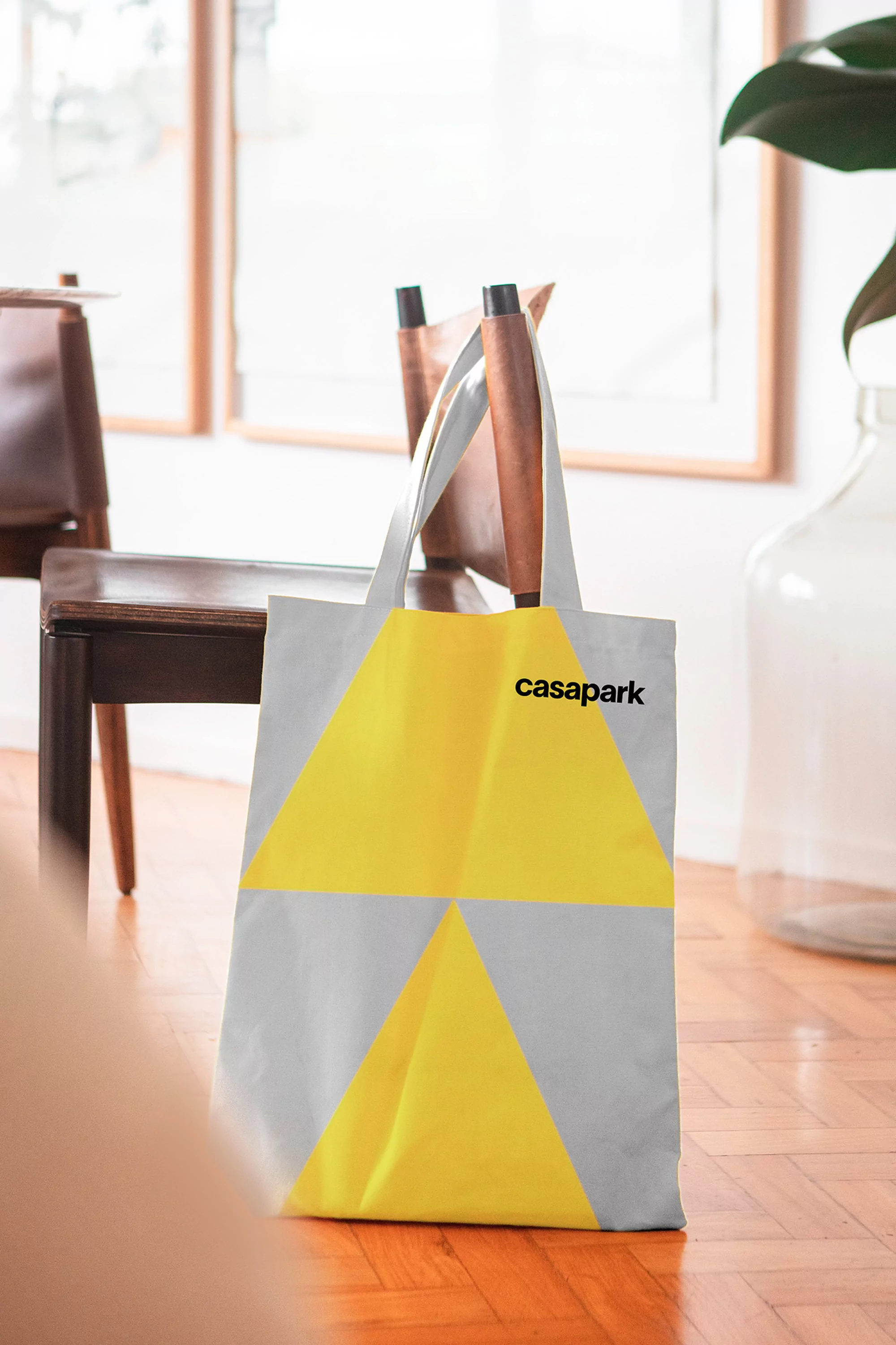 Tote bag with a yellow triangular pattern standing on a wood floor near a wooden chair in an indoor setting with natural light.