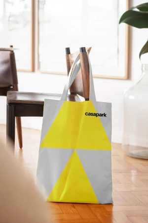 Tote bag with yellow and grey geometric pattern and Casapark logo resting on a wooden parquet floor in a bright room.