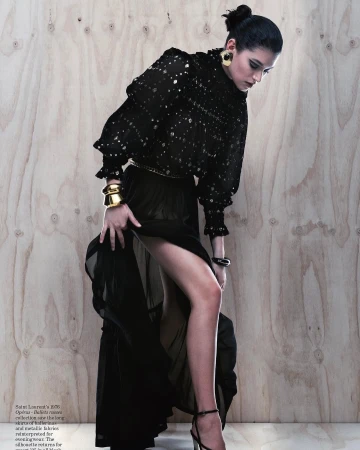 Model in a black sheer blouse and long black skirt lifting her skirt slightly against a light wood panel background.