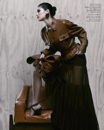 Model wearing brown leather jacket and long pleated skirt holding a large leather bag, posed against a wooden plywood wall.