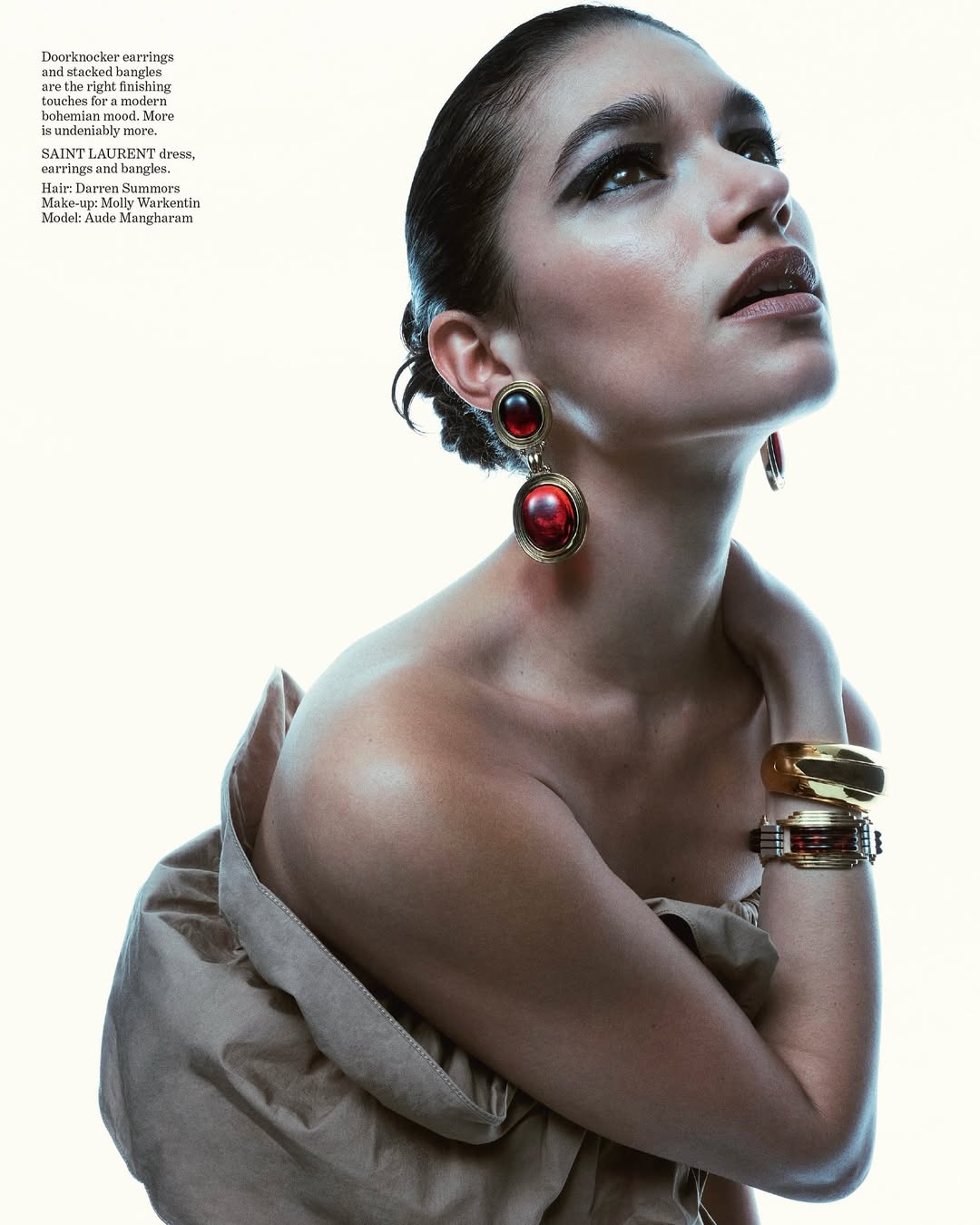 Woman poses in a fashion shot wearing large red and gold earrings, a dress, and gold bracelets against a light background.