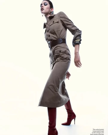 A stylish adult female model in a tan safari-style dress with a wide belt and knee-high red boots strikes a pose against a bright background.