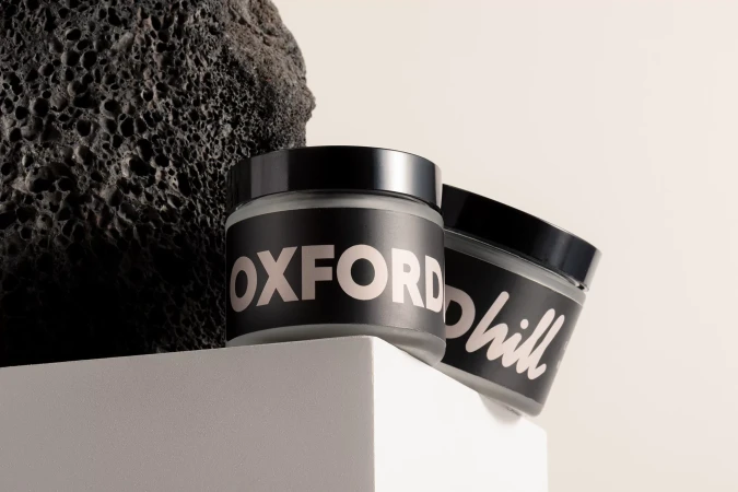 Two cosmetic jars with black labels and lids are displayed against a textured black rock and white platform in a studio setting.