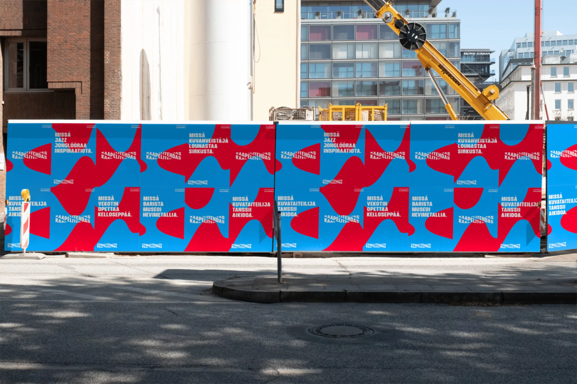 Construction site hoarding featuring red and blue graphic art panels with text and shapes against a bright building and construction crane.