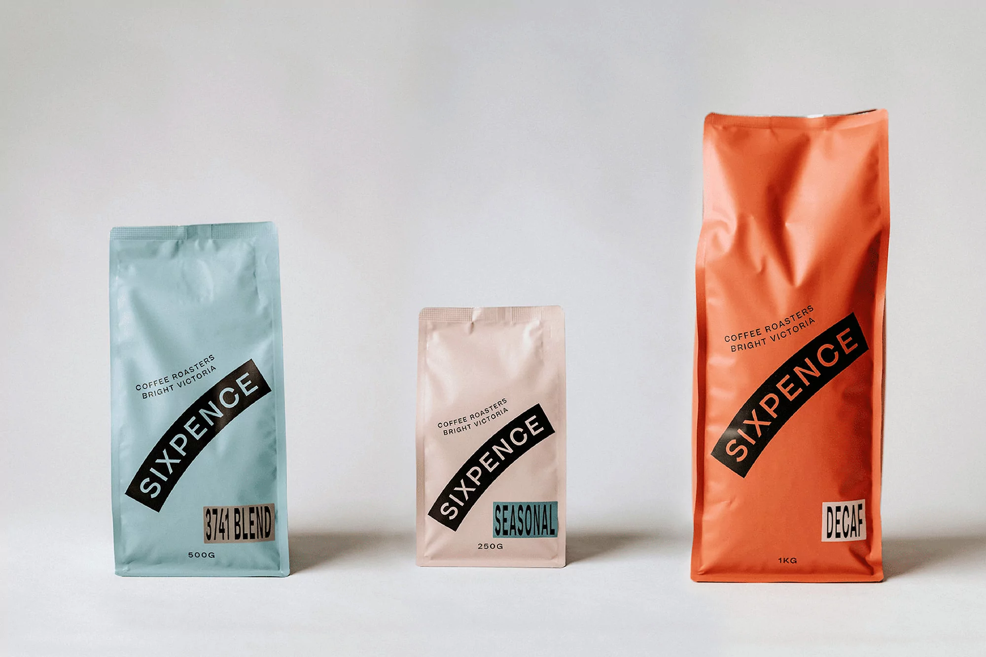 Three colorful Sixpence coffee bags of various sizes displayed on a white surface with a soft studio lighting setup, showcasing product design.