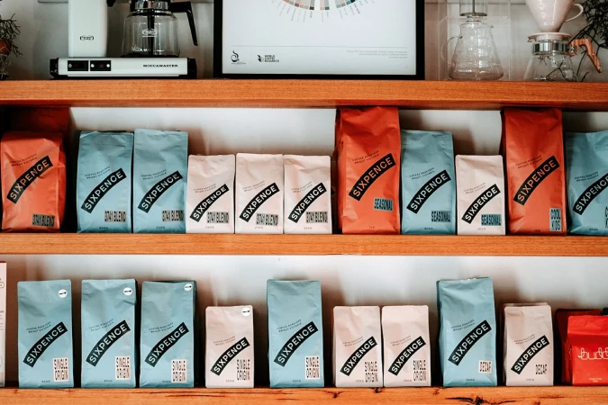 Shelves filled with neatly arranged packages of Sixpence Coffee Roasters coffee beans in various colors and blends, with brewing equipment.