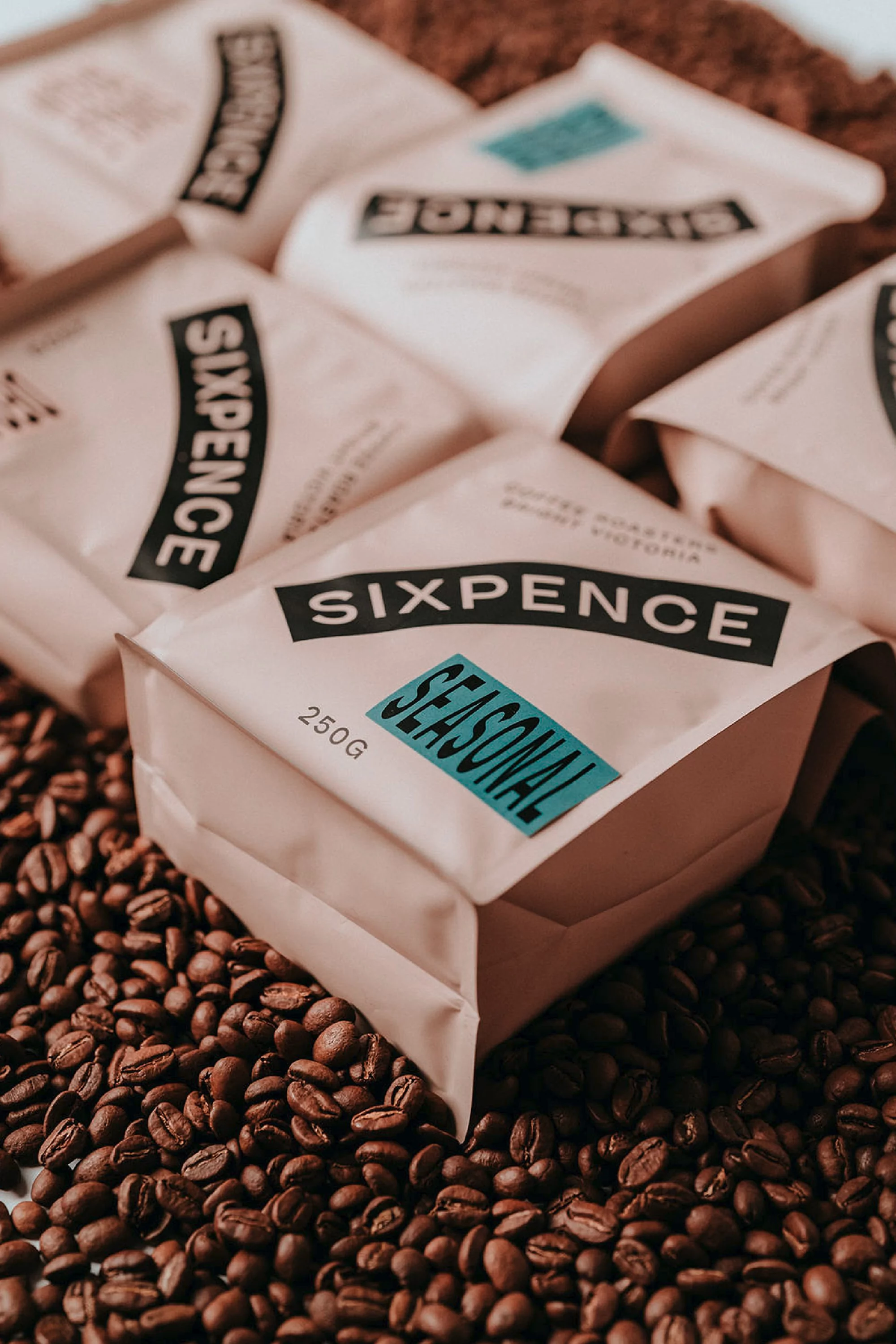 Several beige coffee bean bags with black and teal labels are scattered among loose, roasted coffee beans in an indoor setting.