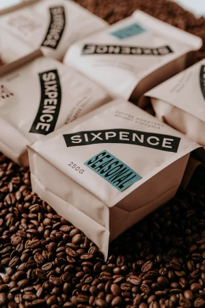 Multiple Sixpence Coffee bags surrounded by scattered roasted coffee beans on a surface.
