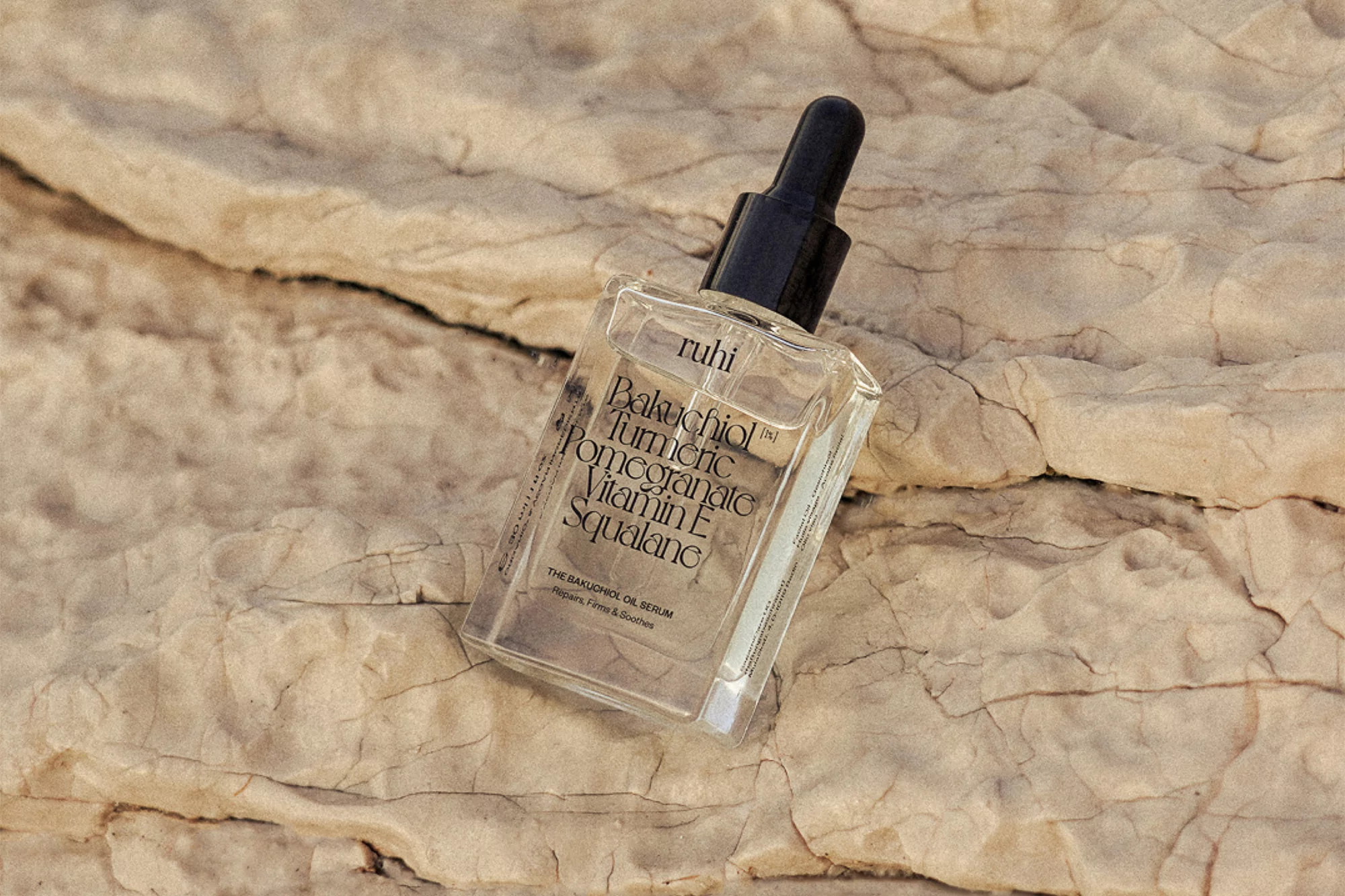 Product shot of a clear glass dropper bottle with black accents, placed diagonally on a textured light-beige rock surface outdoors.