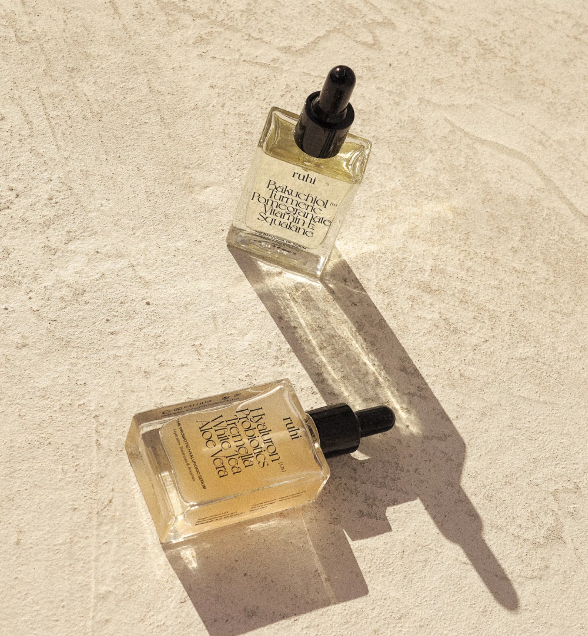 Two square glass bottles of skin serum with black dropper caps sit on a textured light-beige surface in bright, diffused sunlight.