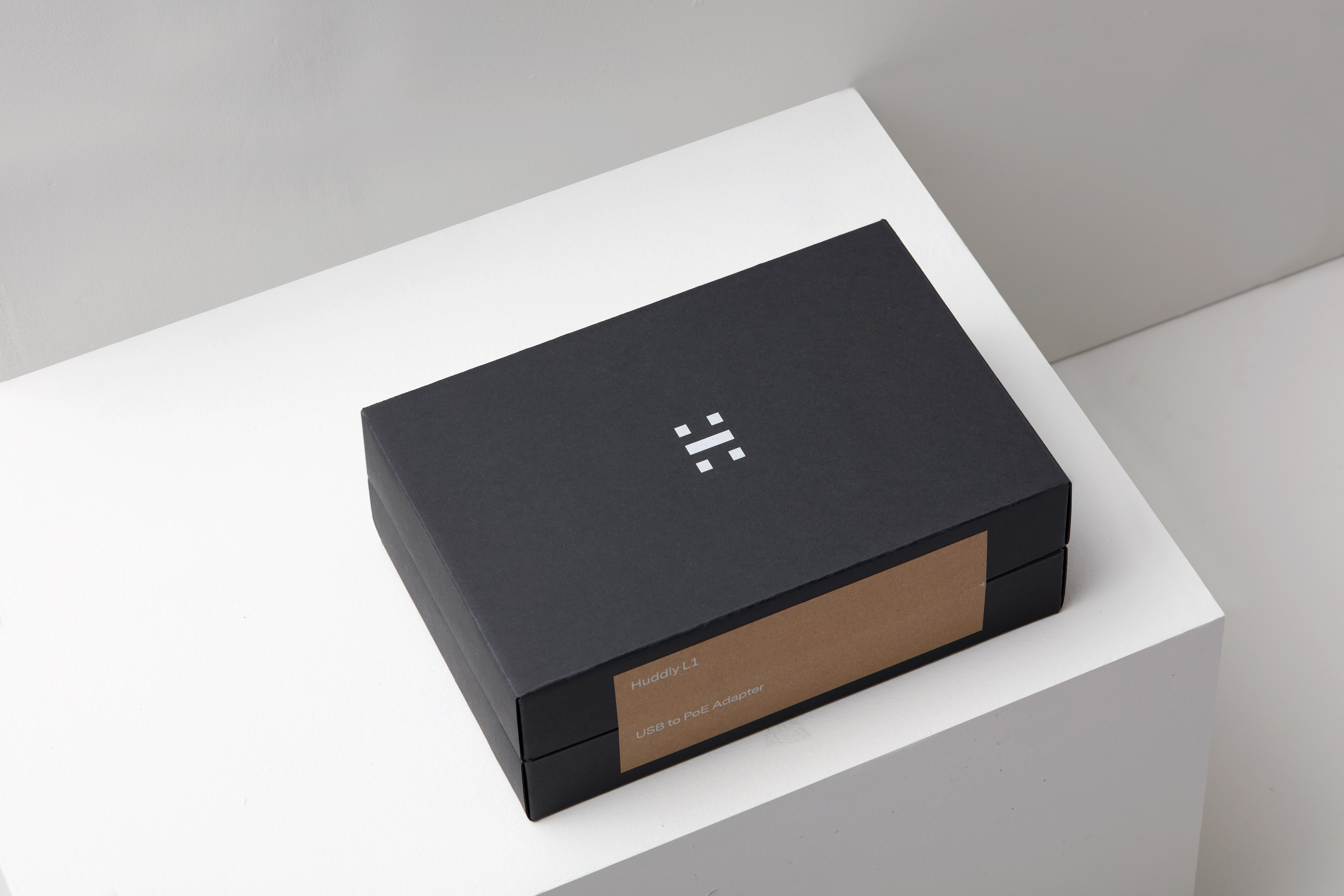 Close-up of a black Huddy L3 adapter box with white logo and brown label resting on a minimalist white platform.