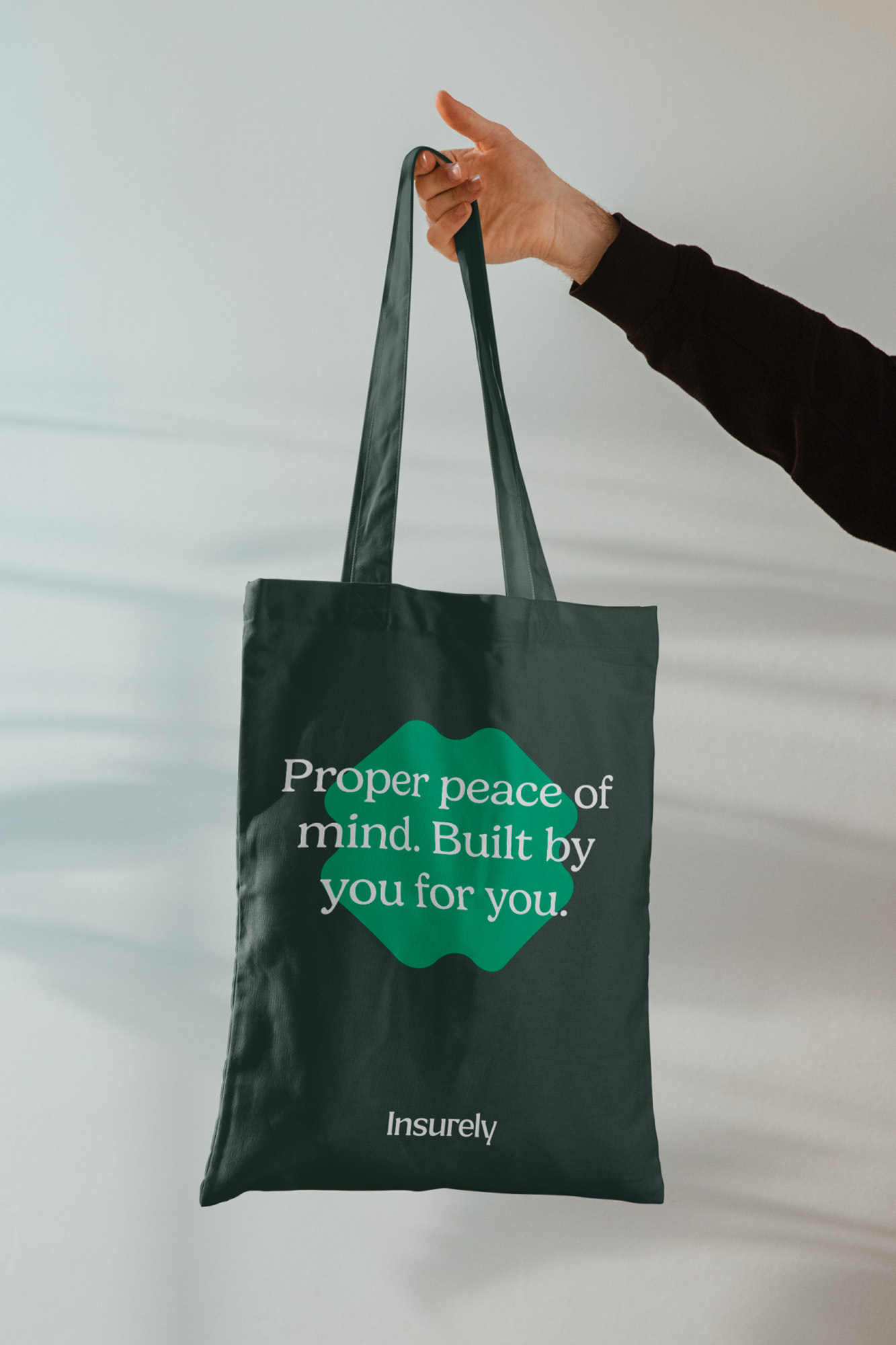 A dark green tote bag with an insurance company logo is held up by a person against a white background in a product-focused studio shot.