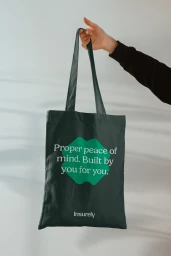 Hand holding a dark green canvas tote bag with white text against a plain light wall