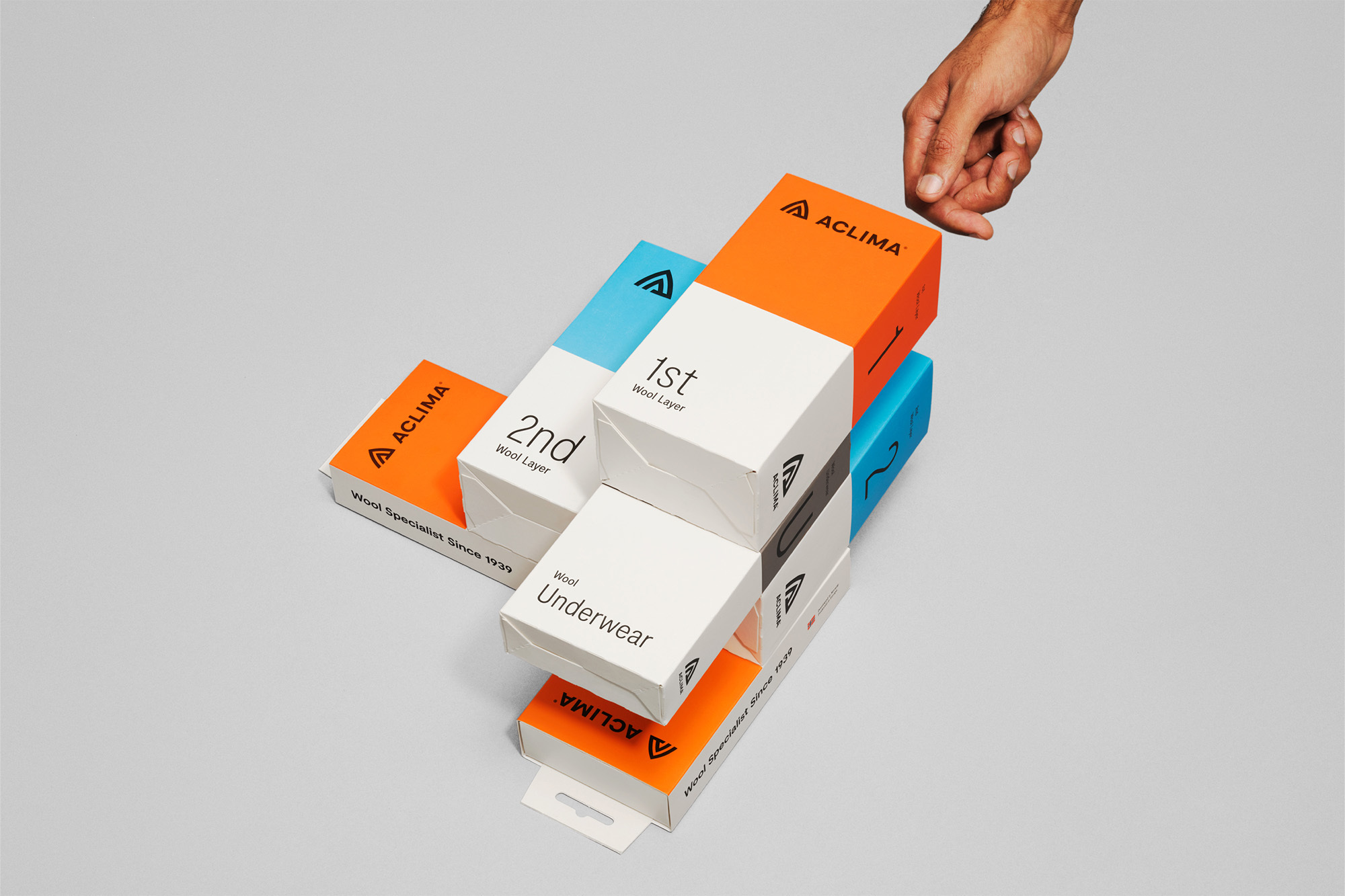 Stacked boxes of ACLIMA wool layers, including the 1st, 2nd, and Wool Underwear boxes, with a hand reaching in, neutral background, product shot.