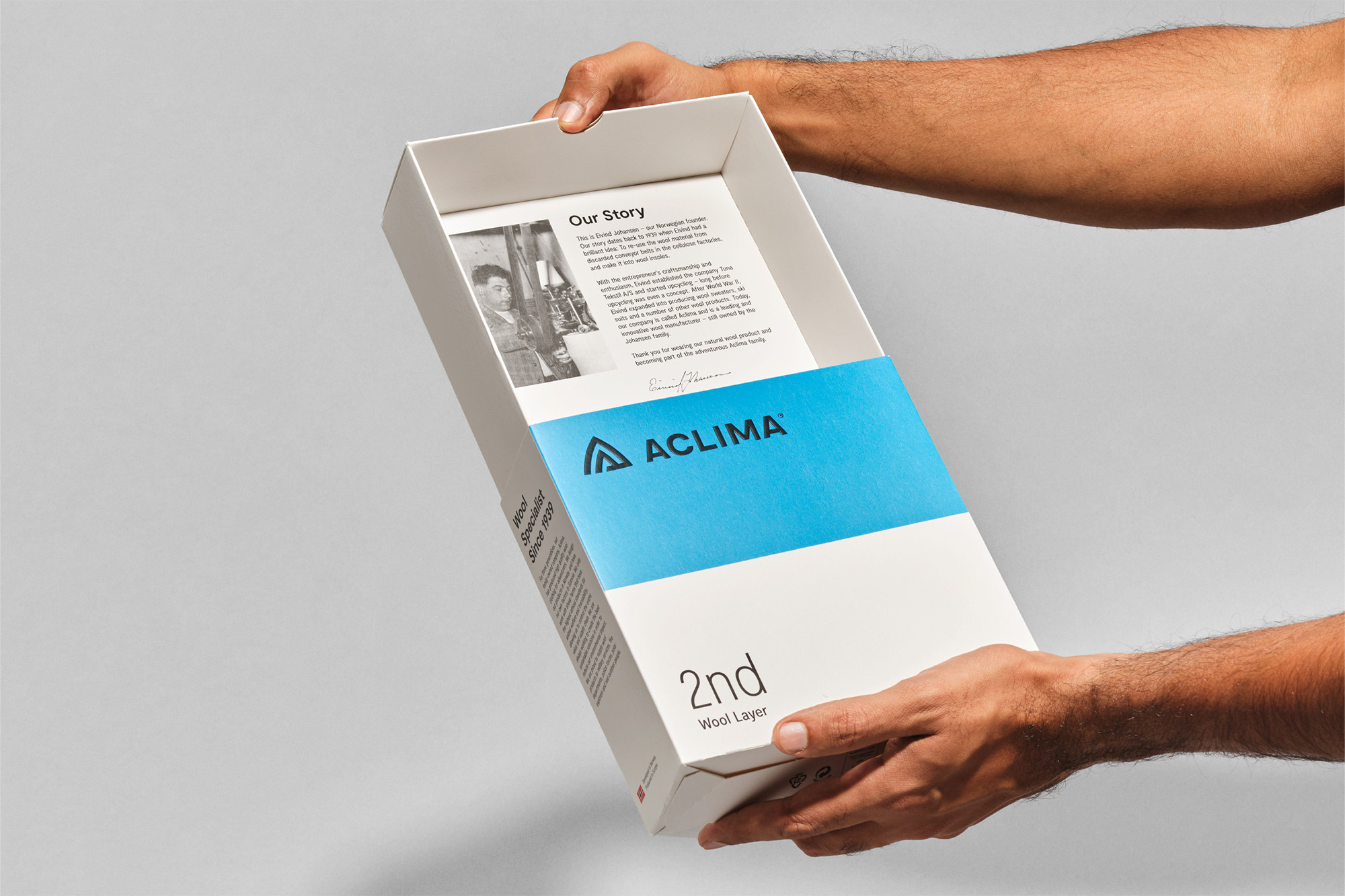 Hands holding a white box with a blue band, featuring the 'Aclima' logo and text about wool layers against a neutral background.