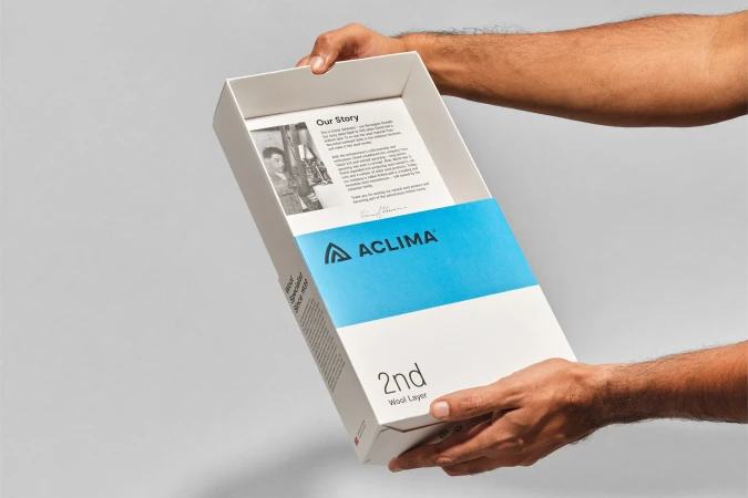 Hands holding a white box with a blue band, featuring the 'Aclima' logo and text about wool layers against a neutral background.
