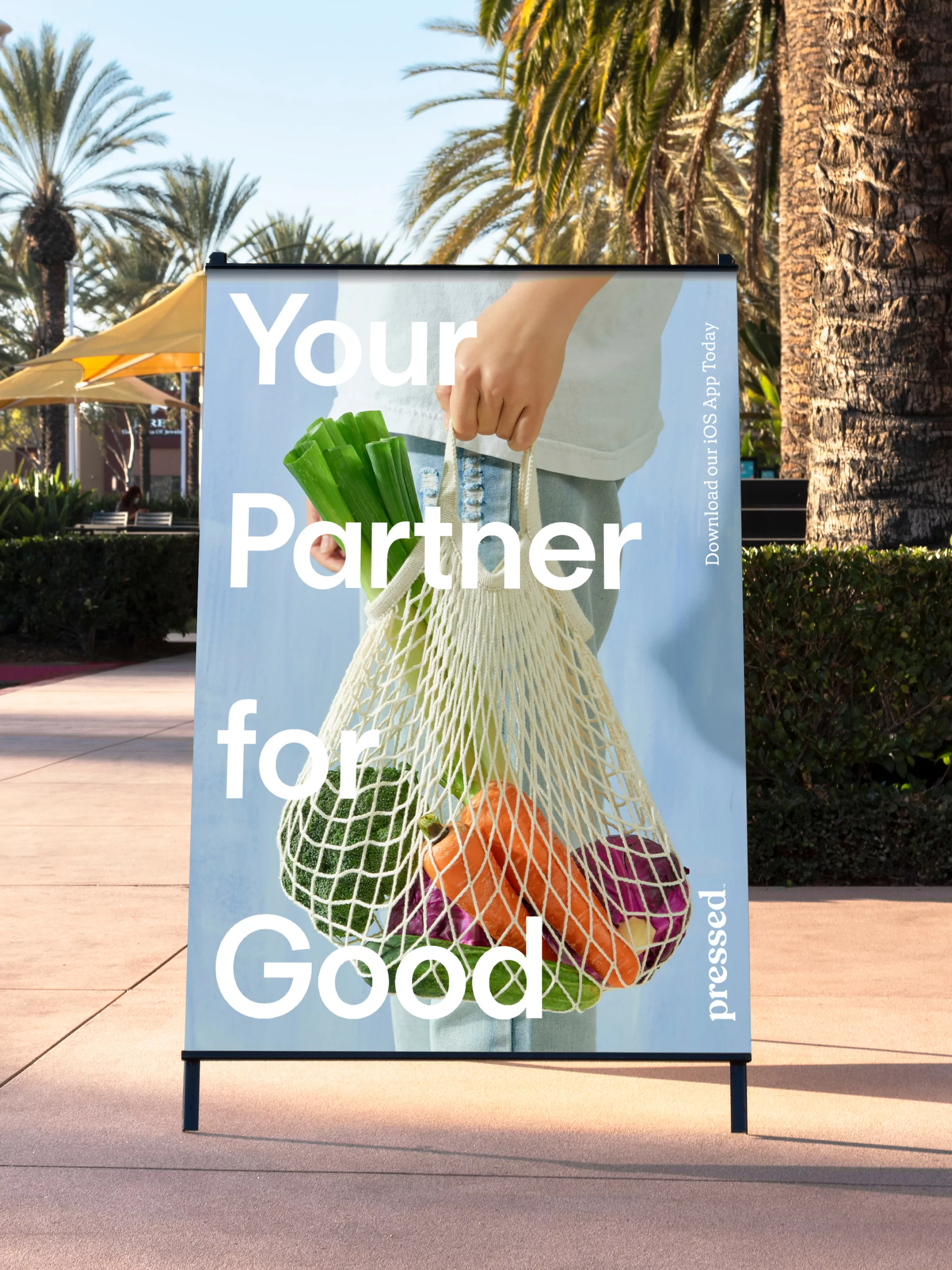 Advertisement for Pressed juice featuring a person holding a string bag with produce, displayed outdoors with palm trees.