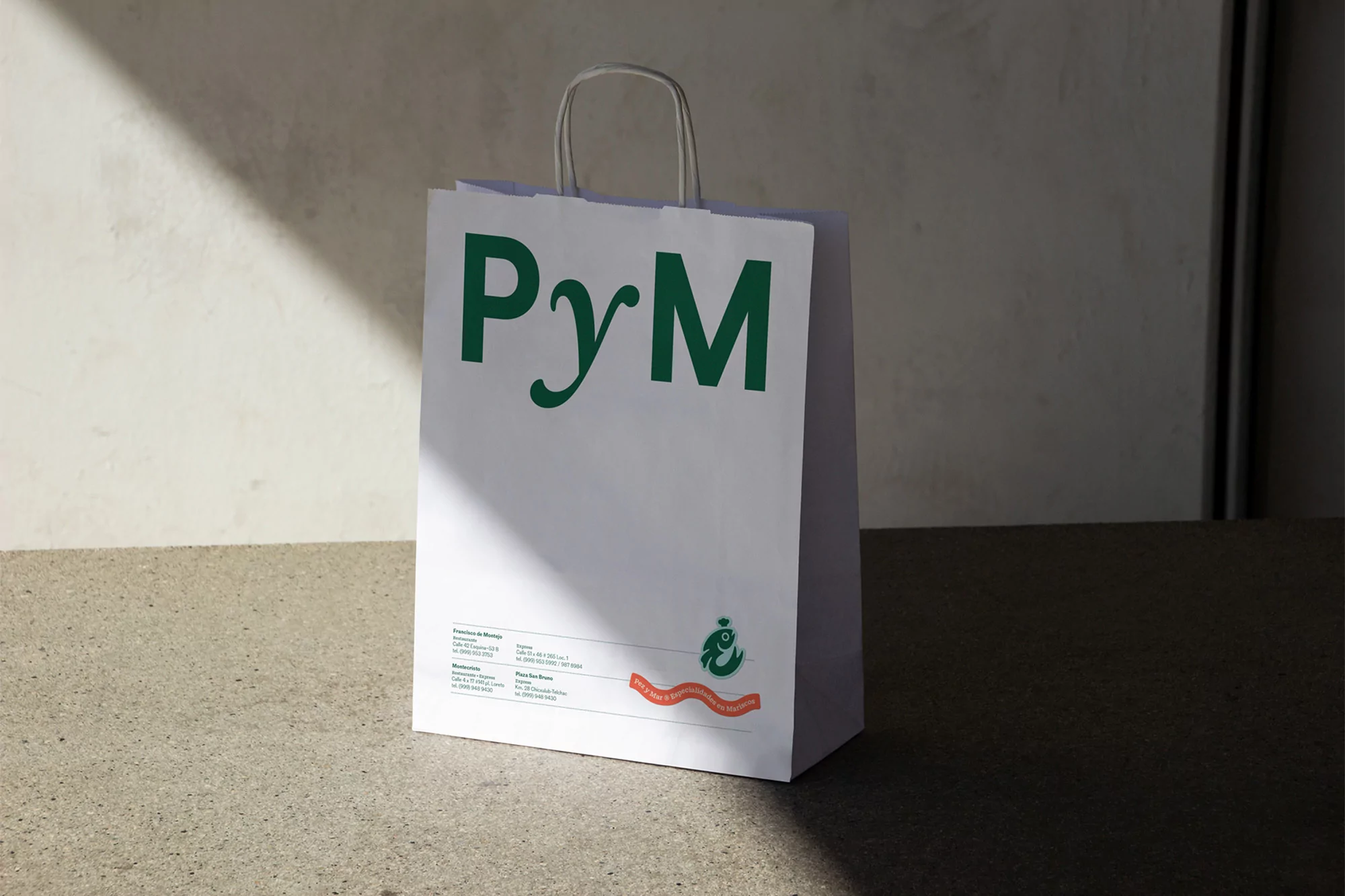 A white paper shopping bag with green text sits on a concrete surface against a light gray wall with sunlight casting shadows.