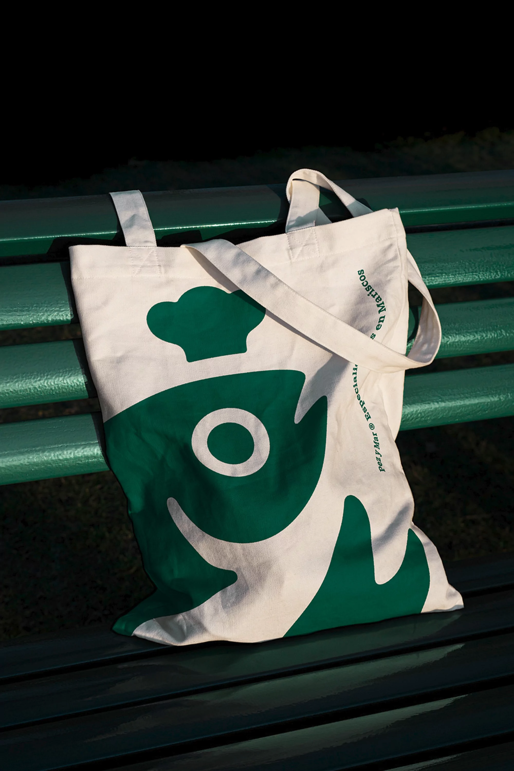 Canvas tote bag on a green park bench features a dark green graphic of a fish wearing a chef's hat in outdoor setting.