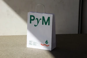 White paper shopping bag with green PyM logo and twisted paper handles standing on a dark stone surface.