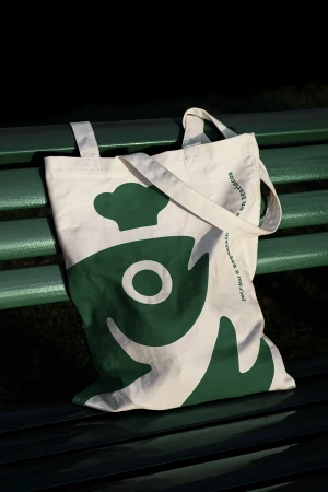 White canvas tote bag with bold green fish graphic resting on a dark green wooden park bench outdoors.