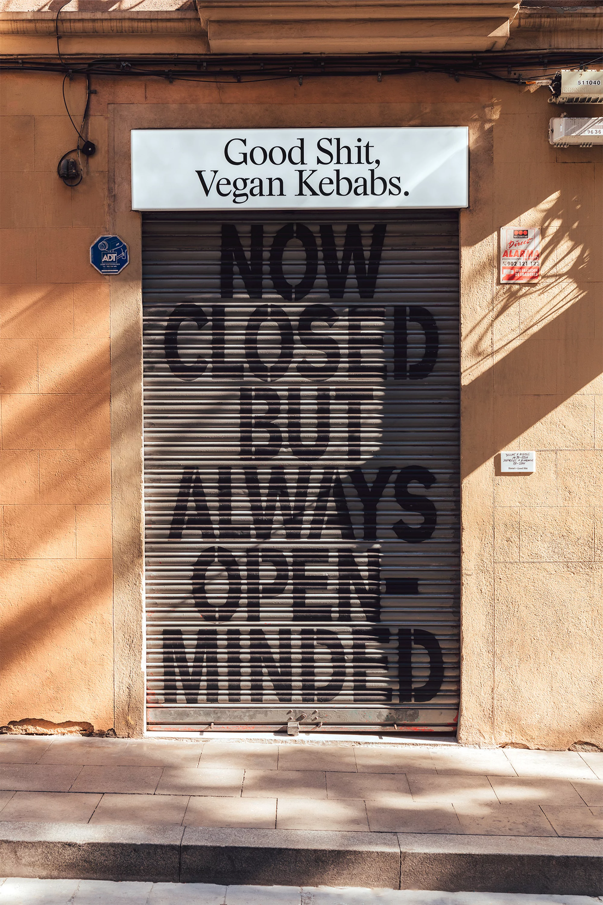 A building with a closed shutter that says "Now Closed But Always Open Minded" with a sign above that says "Good Shit, Vegan Kebabs."