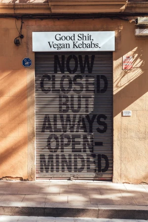 A building with a closed shutter that says "Now Closed But Always Open Minded" with a sign above that says "Good Shit, Vegan Kebabs."