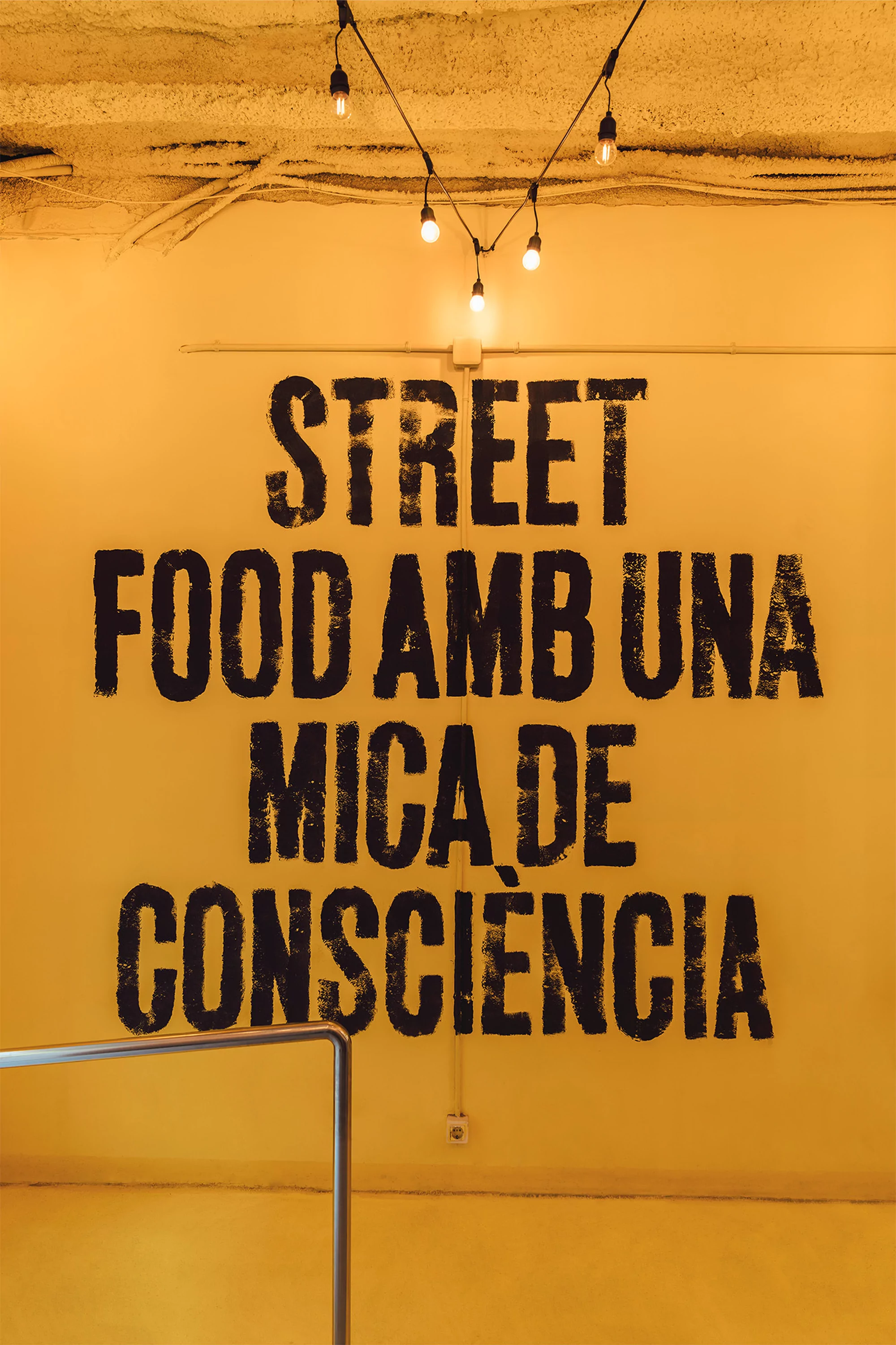 Yellow wall with 'Street Food Amb Una Mica De Consciencia' in black text, lit by string lights, creating an urban, food-focused interior.