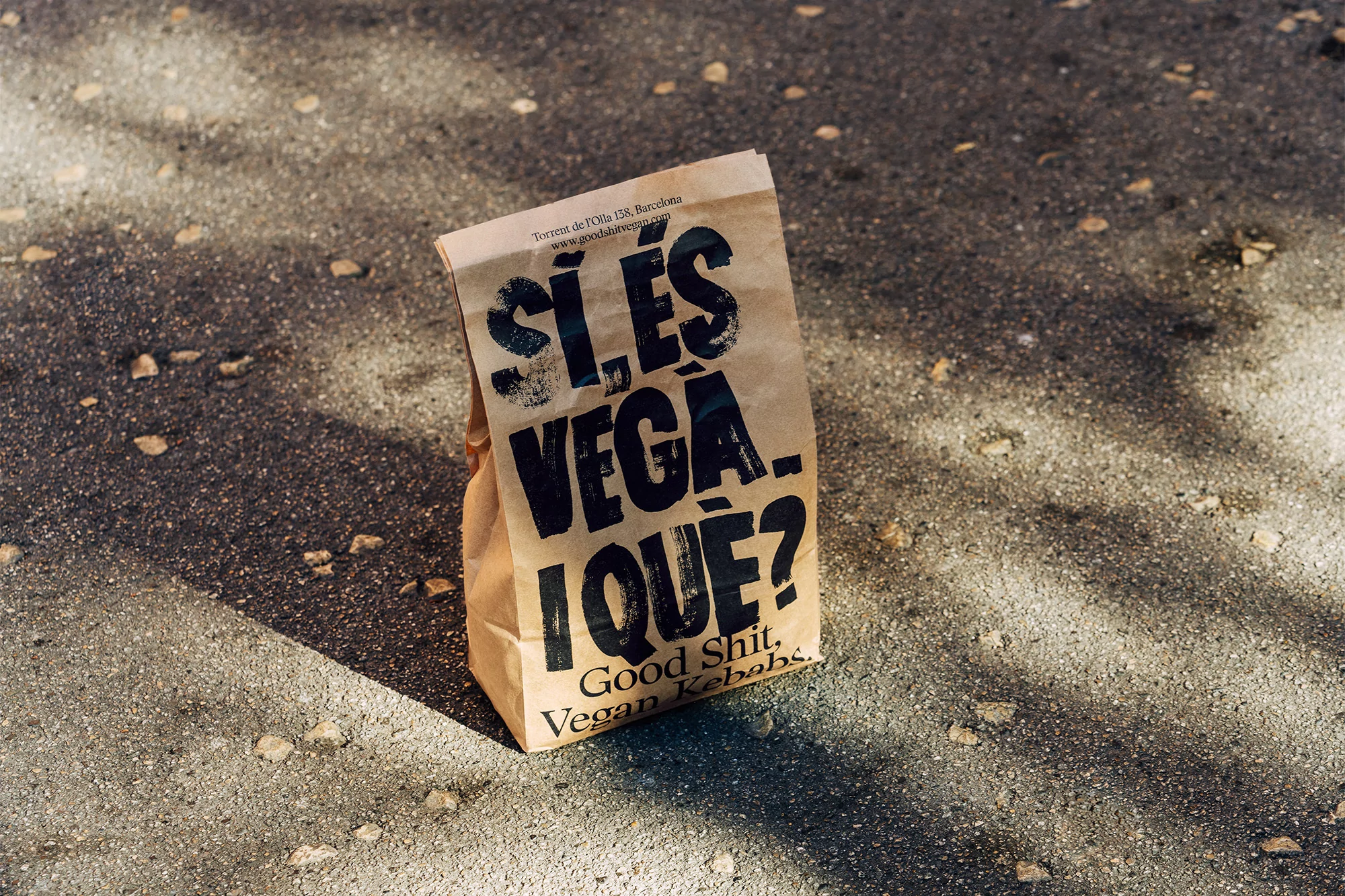 Brown paper bag with bold text 'Sí. ES VEGA. IQUE?' and 'Good Shit, Vegan Kebabs' on concrete ground in diffused sunlight.