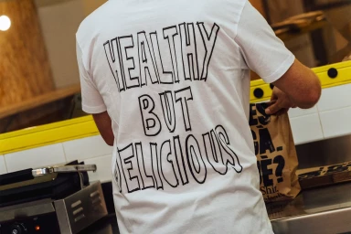Person wearing a white t-shirt with black typographic text reading HEALTHY BUT DELICIOUS while packing a brown paper bag