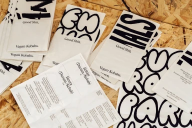 A flat lay of branded paper packaging, menus, and stickers for Good Shit Vegan Kebabs on an OSB wood background.