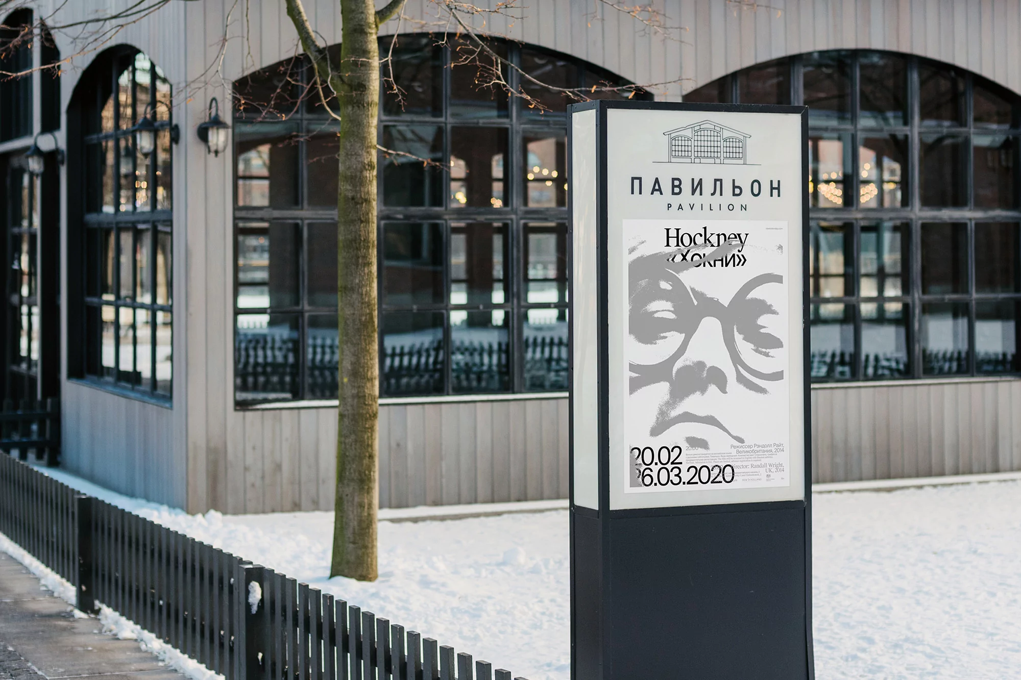 Outdoor shot of a poster for Hockney exhibition next to a building with large windows and snow in the foreground on a winter day.