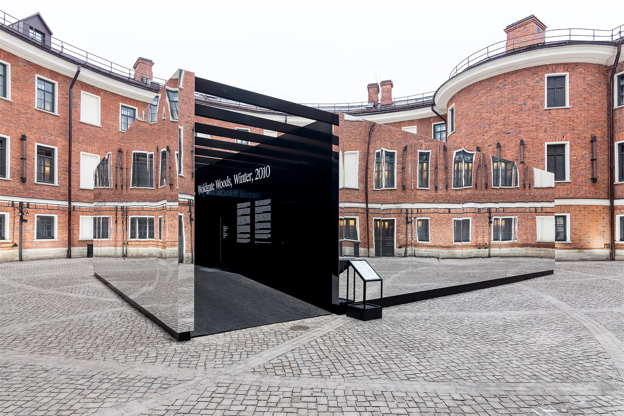 Courtyard featuring a modern art installation with black panels and mirror-like surfaces reflecting a red brick building.