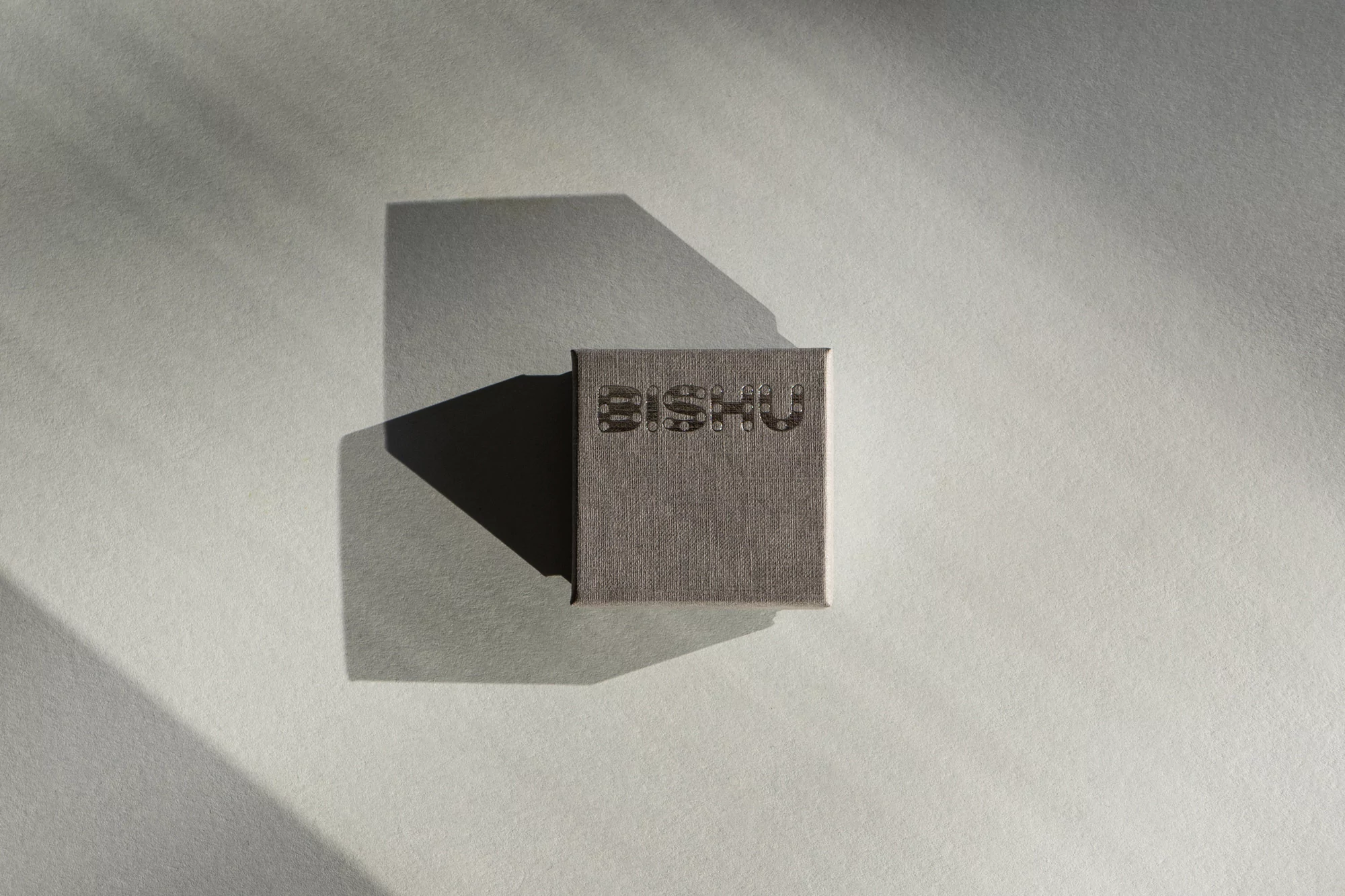 Close-up of a gray jewelry box with the word "BISHU" on top, centered on a light gray surface, with shadow on the left side.