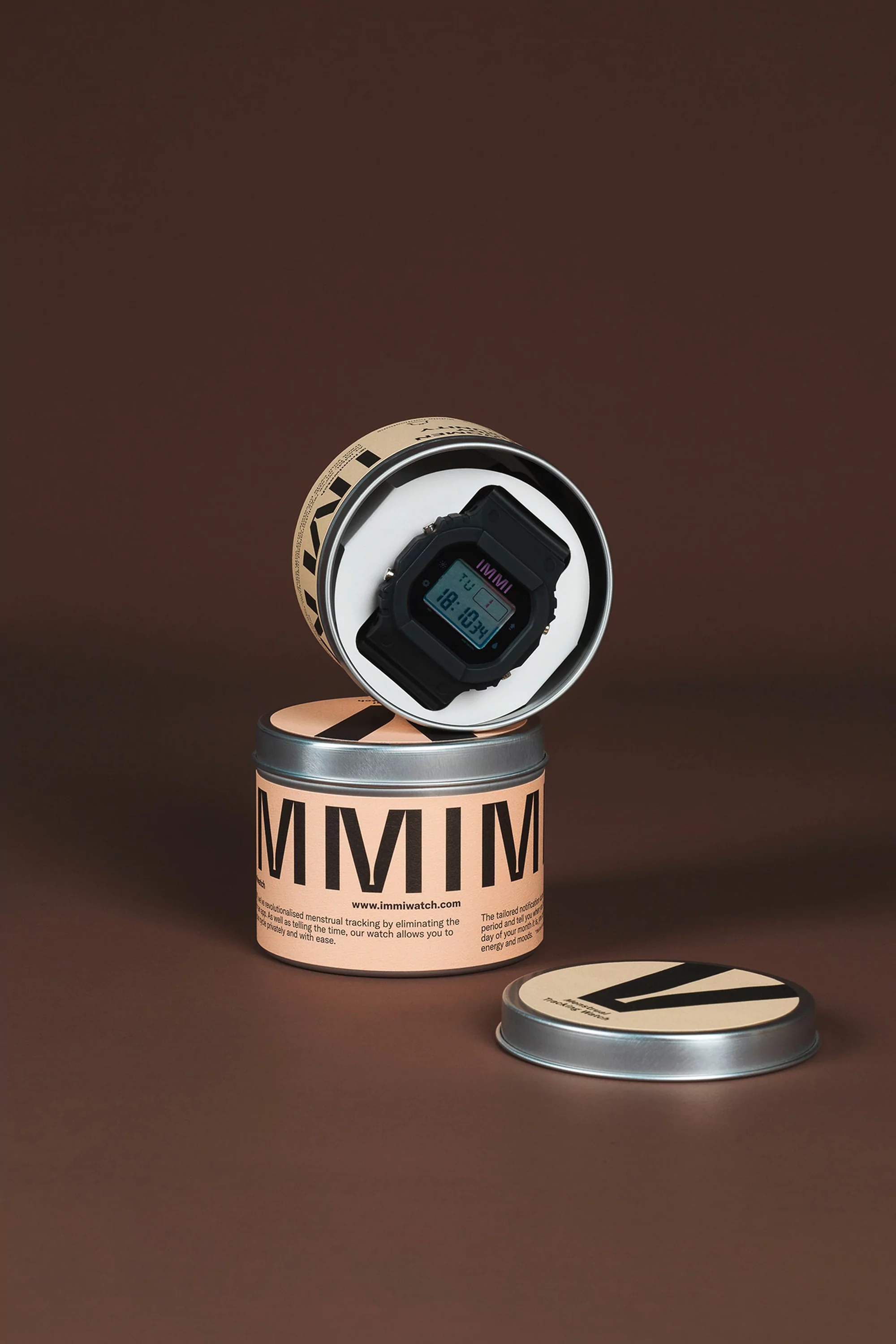 Black digital watch displayed on and in a branded metal tin. The watch face shows the time in digital numbers on a neutral background.