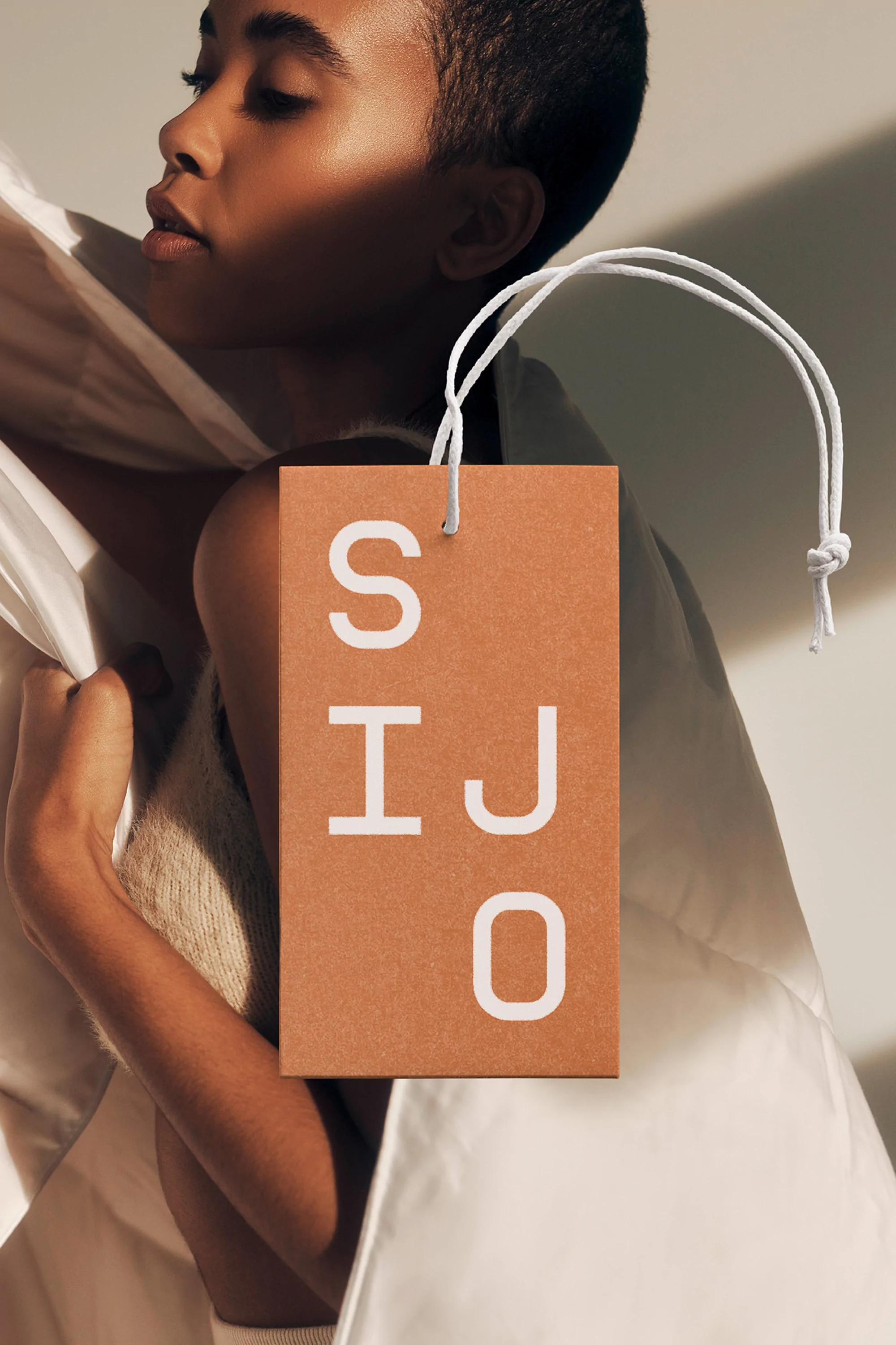 Close-up of a young Black woman draped in white fabric, face partially visible, holding up a brown tag with 'SHJO' printed on it.
